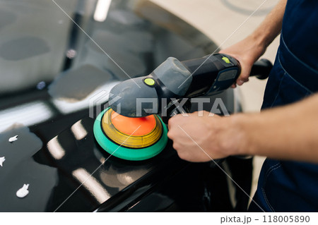 Closeup top view of unrecognizable mechanic male in protective gloves polishing surface of hood of black car using orbital polisher in repair shop. Concept of professional car repair, maintenance work 118005890