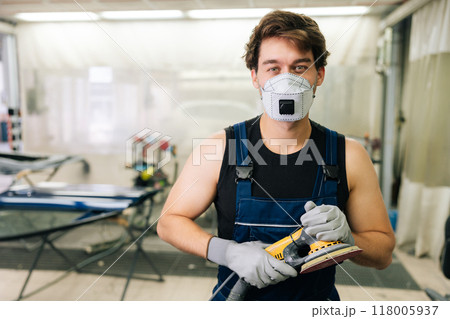 Mechanic male in protective mask holding machinery car polishers maintenance to remove marks repair. Portrait of car paint technician in uniform holding car polisher to polish of car with refinement. 118005937