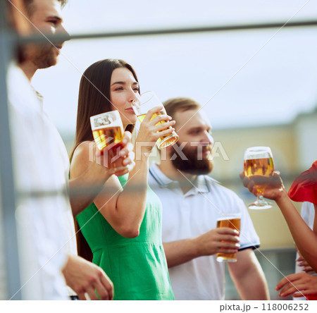 Group of friends enjoys sunny day together, sipping cold beers. Woman in green dress takes drink, surrounded by smiling faces and relaxed vibe. 118006252