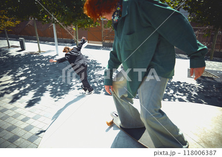 Energy and extreme. Dynamic image of two men, young guys in motion on skateboard, showing skills and trick in public park. Energy and extreme. Dynamic image of two men, young guys in motion on skateboard, showing skills and trick in public park. 118006387