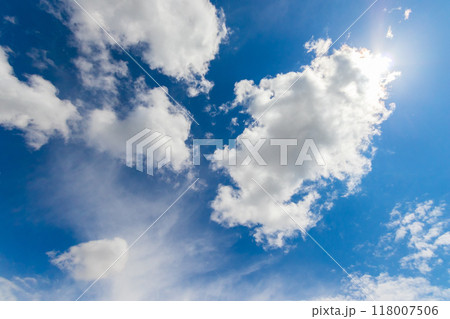 Blue sky is adorned with fluffy white cumulus clouds, allowing the sun to shine through. A natural sky landscape resembling a painting of meteorological phenomenon. 118007506