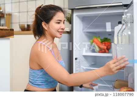 Smiling young woman dressed in casual workout attire reaching for something inside the fridge, preparing for healthy meal Smiling young woman dressed in casual workout attire reaching for something inside the fridge, preparing for healthy meal 118007790
