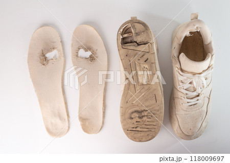Old, worn-out white sneaker with partially detached sole and missing insole. Visible dirt, wear, and holes in the shoe. Removed sole and insole are lying next to it, 118009697