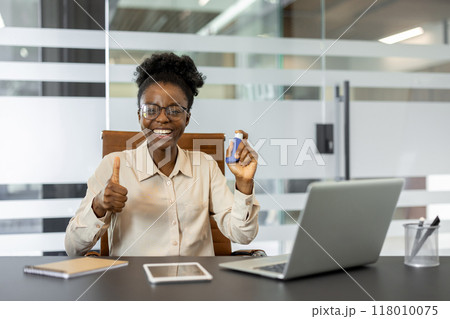 African American businesswoman in office smiling with confidence, holding inhaler and giving thumbs up. Surroundings include laptop, tablet, and phone, creating modern professional setting. 118010075