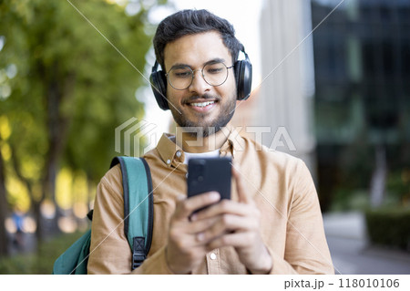 Young man with headphones using smartphone outdoors. Enjoying music, wearing casual attire with backpack. Displaying relaxation, enjoying day. Captures lifestyle, modern technology, student life. Young man with headphones using smartphone outdoors. Enjoying music, wearing casual attire with backpack. Displaying relaxation, enjoying day. Captures lifestyle, modern technology, student life. 118010106