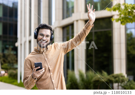 Cheerful man wearing headphones smiling while holding phone and waving outside modern building. Person enjoying music and technology in casual setting, conveying happiness and connection in outdoor 118010133