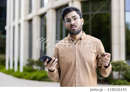 Man with glasses standing outside, holding phone and credit card, showing confused expression. Concept of financial uncertainty, technology frustration, decision making, or communication issues. Man with glasses standing outside, holding phone and credit card, showing confused expression. Concept of financial uncertainty, technology frustration, decision making, or communication issues. 118010145