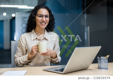 Confident businesswoman in contemporary office setting relaxing with coffee. She sits at desk using laptop, smiling pleasantly. Represents workplace comfort, professional success environment Confident businesswoman in contemporary office setting relaxing with coffee. She sits at desk using laptop, smiling pleasantly. Represents workplace comfort, professional success environment 118010374