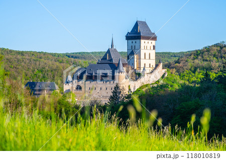 The majestic Karlstejn Castle stands proud against a backdrop of lush greenery as the morning sun rays enhance its architecture. A peaceful scene unfolds in this historic location of Czechia. The majestic Karlstejn Castle stands proud against a backdrop of lush greenery as the morning sun rays enhance its architecture. A peaceful scene unfolds in this historic location of Czechia. 118010819