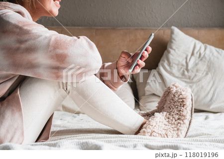 Woman in cozy attire using smartphone while sitting on bed. Woman in cozy attire using smartphone while sitting on bed. 118019196