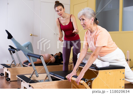 Trainer controlling movements of senior woman doing pilates on combo chair Trainer controlling movements of senior woman doing pilates on combo chair 118020634