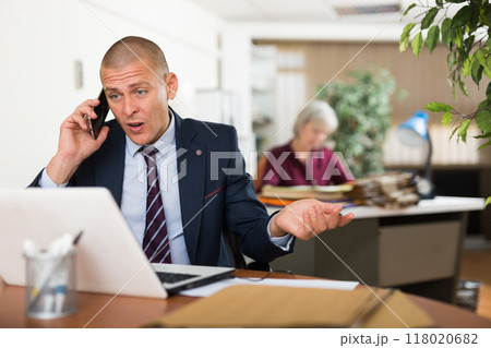 Irritated businessman talking on phone and working with laptop in office Irritated businessman talking on phone and working with laptop in office 118020682