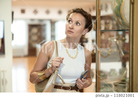 Woman with piece of paper and phone in hands walks around museum hall, view exhibition 118021001