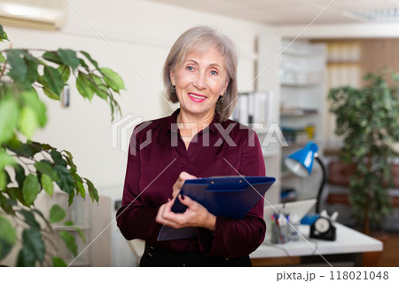 Friendly aged female administrative secretary standing in office with folder of documents Friendly aged female administrative secretary standing in office with folder of documents 118021048