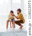 Mother cuts her son's hair by herself. Little boy sits, covered with cloth, and holds pair of scissors. Beauty at home lifestyle. 118022073