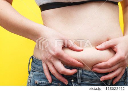 Woman hands compresses the fat on her belly on a yellow background. 118022462