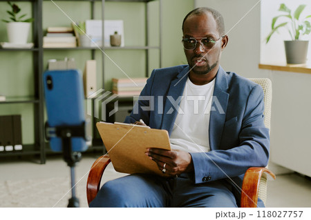 Portrait of businessman holding clipboard while sitting in modern office with shelves, plant and phone on tripod in background capturing video 118027757