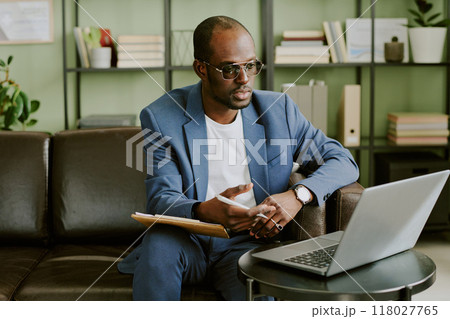 Man in blue suit and glasses conducting business meeting online while sitting on brown leather sofa in modern office with green walls and shelves behind Man in blue suit and glasses conducting business meeting online while sitting on brown leather sofa in modern office with green walls and shelves behind 118027765