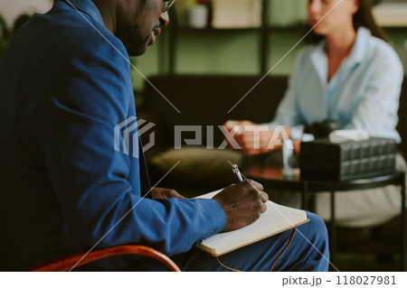 Black man in a blue suit taking notes during a meeting, with a blurred background of a woman seated at table in a casual setting, indicating a professional environment Black man in a blue suit taking notes during a meeting, with a blurred background of a woman seated at table in a casual setting, indicating a professional environment 118027981