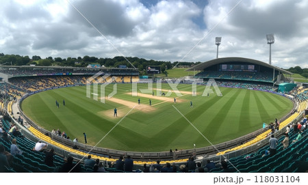 Wide view of cricket stadium during match. Cricket match in progress at a stadium with a lush green pitch, players in action, and a crowd watching from the stands. 118031104