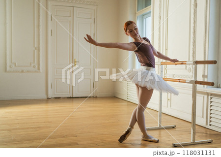 Young ballerina in tutu skirt stretching practicing ballet positions dancing near ballet barre in dance class. Young classical dancer woman in dance studio. Ballet class training Ballet school concept 118031131