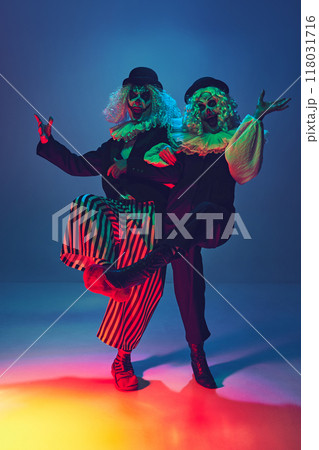Crazy, frightening clowns with furious make-up dressed vintage attire dancing in neon light against gradient blue background. 118031716