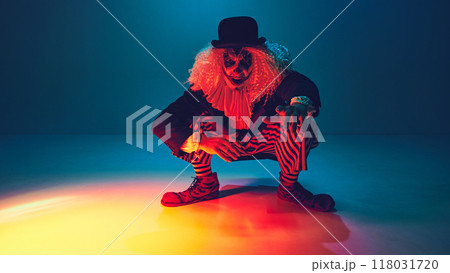 Crouched with menace, clown dressed retro clothes reaches forward, in neon light against gradient background. 118031720