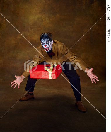 Clown with spooky make-up trying to catch red present with shocked expression against vintage background. Clown with spooky make-up trying to catch red present with shocked expression against vintage background. 118031757