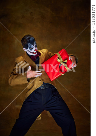 Holding gift tightly, clown looks excited yet sinister, as if hiding twisted surprise inside against vintage dark background. 118031771