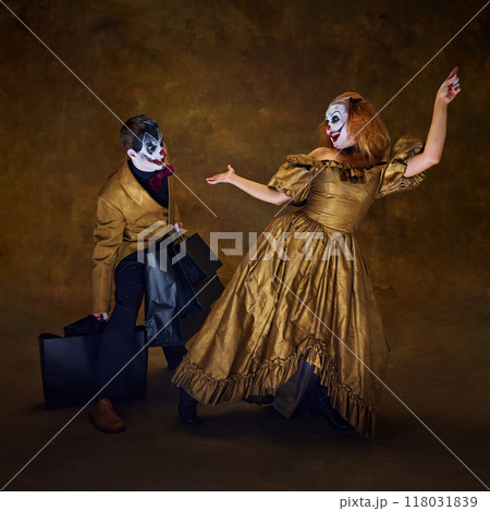 Two clowns with frightening make-up, one bending under weight of black shopping bags, while other poses against vintage backdrop. 118031839
