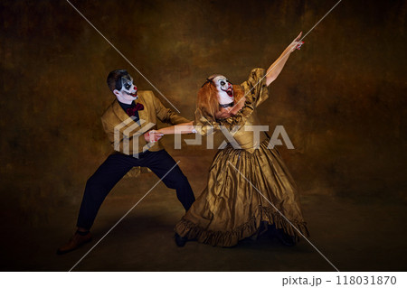 Clown duo, pulling off exaggerated dramatic movements, one pointing in distance while other holds her back against vintage style backdrop. Clown duo, pulling off exaggerated dramatic movements, one pointing in distance while other holds her back against vintage style backdrop. 118031870