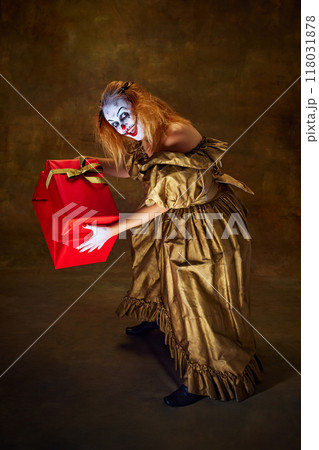 Woman with spooky make-up in vintage gown, bending down playfully while clutching large gift box against vintage style backdrop. Woman with spooky make-up in vintage gown, bending down playfully while clutching large gift box against vintage style backdrop. 118031878