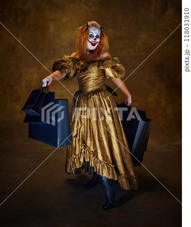 Horror-clown woman dressed in antique-style gown, holding large shopping bags, smiling wickedly, posing against vintage backdrop. Horror-clown woman dressed in antique-style gown, holding large shopping bags, smiling wickedly, posing against vintage backdrop. 118031910