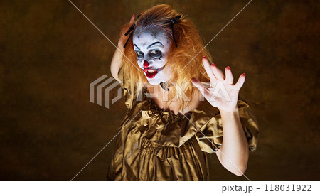 Frightening clown woman in golden gown, raising her hands with wild expression, posing dramatically against vintage background. 118031922