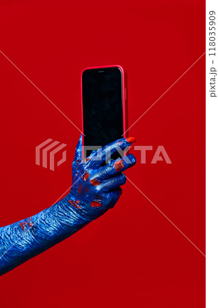Zombie's, monster's blue hand holds smartphone with blank screen against red studio background. Zombie's, monster's blue hand holds smartphone with blank screen against red studio background. 118035909