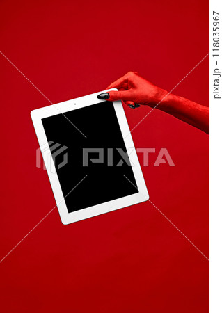 Horror red devil's hand with blank long nails holding tablet with blank screen against red studio background. Horror red devil's hand with blank long nails holding tablet with blank screen against red studio background. 118035967