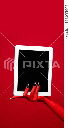 Horror red devil's hand with blank long nails holding tablet with blank screen against red studio background. Horror red devil's hand with blank long nails holding tablet with blank screen against red studio background. 118035968