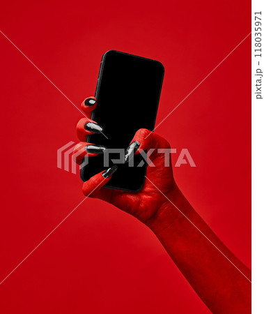 Horror red devil's hand with blank long nails holding smartphone with blank screen against red studio background. Horror red devil's hand with blank long nails holding smartphone with blank screen against red studio background. 118035971