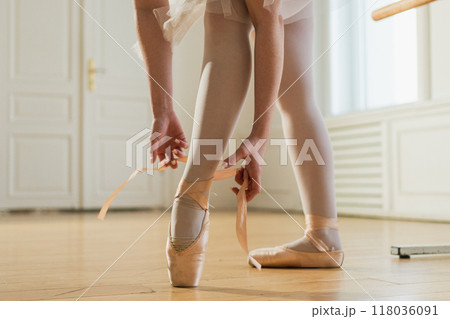 Ballerina puts on pointe shoes on leg in dance class. Classical ballet dancer woman in dance studio. Ballerina putting on tying classical pointe shoes for dance training. Ballet school concept 118036091