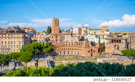 The ancient Trajan Market stands proudly in Rome, Italy, showcasing its remarkable architecture while surrounded by blooming greenery and majestic ancient ruins under a bright blue sky. 118036527