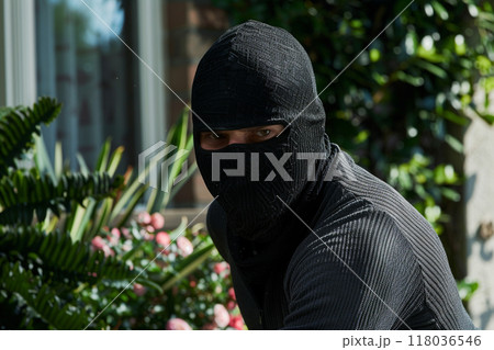 A masked burglar cautiously surveying the surroundings in a garden near a house, with greenery in the background. A masked burglar cautiously surveying the surroundings in a garden near a house, with greenery in the background. 118036546