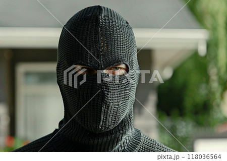 A masked criminal standing in front of a house, wearing a black balaclava and a textured outfit. 118036564