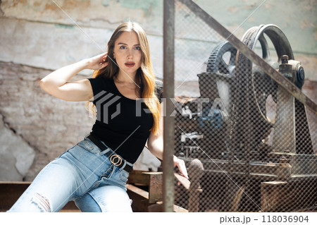 Woman in black t-shirt and jeans standing near wire fencing in abandoned factory Woman in black t-shirt and jeans standing near wire fencing in abandoned factory 118036904