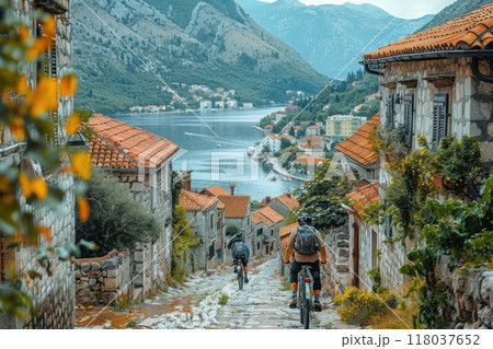 Two bikers ride down a stone street in a small European village with a waterfront. 118037652