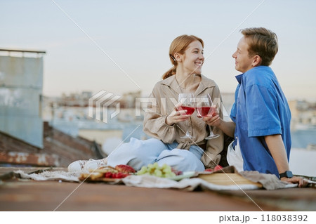 Couple smiling while holding wine glasses and enjoying picnic on rooftop with cityscape backdrop at sunset. Fruits and cheese placed on blanket while sharing laughter 118038392