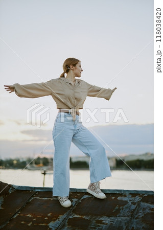 Graceful woman balancing on rooftop with arms outstretched, wearing light attire and looking horizon, scenic sky in background, conveying tranquility and freedom 118038420