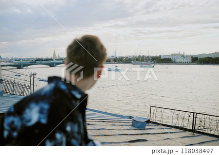Individual with short hair standing on rooftop, facing away from camera and observing cityscape with river, boats, and historic buildings in background 118038497