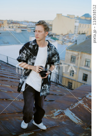 Young man standing on rooftop holding camera, with cityscape in background looking thoughtful. Casual apparel and urban surroundings with sunset lighting atmosphere Young man standing on rooftop holding camera, with cityscape in background looking thoughtful. Casual apparel and urban surroundings with sunset lighting atmosphere 118038512