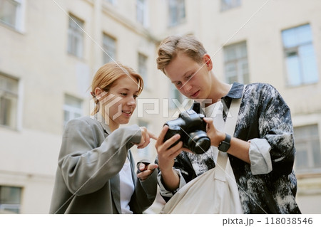 Young individuals examining camera settings in outdoor cityscape, surrounded by buildings, one holding professional camera while other points at screen, showcasing interest in photography Young individuals examining camera settings in outdoor cityscape, surrounded by buildings, one holding professional camera while other points at screen, showcasing interest in photography 118038546