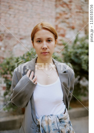 Portrait of Caucasian woman looking at camera while standing against brick wall and greenery background, holding jacket with one hand without smiling 118038556